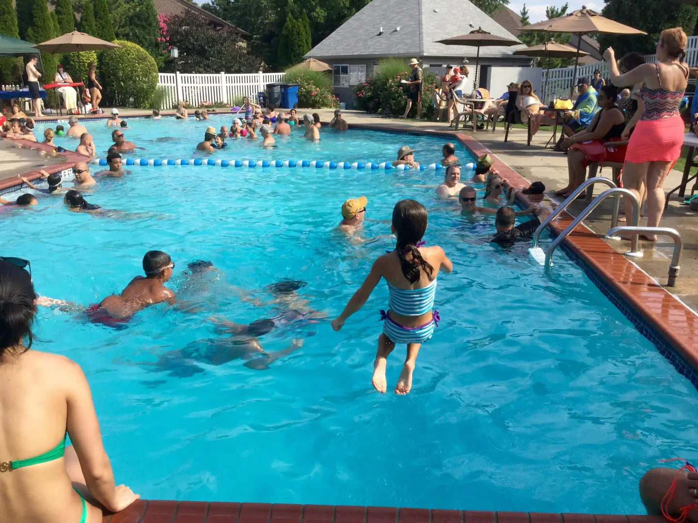 A child jumps into a crowded swimming pool, surrounded by people relaxing and socializing on a sunny day.