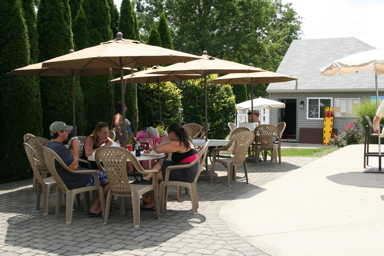 People sitting and dining at outdoor tables under umbrellas on a sunny day, with a small building in the background and trees lining the area.