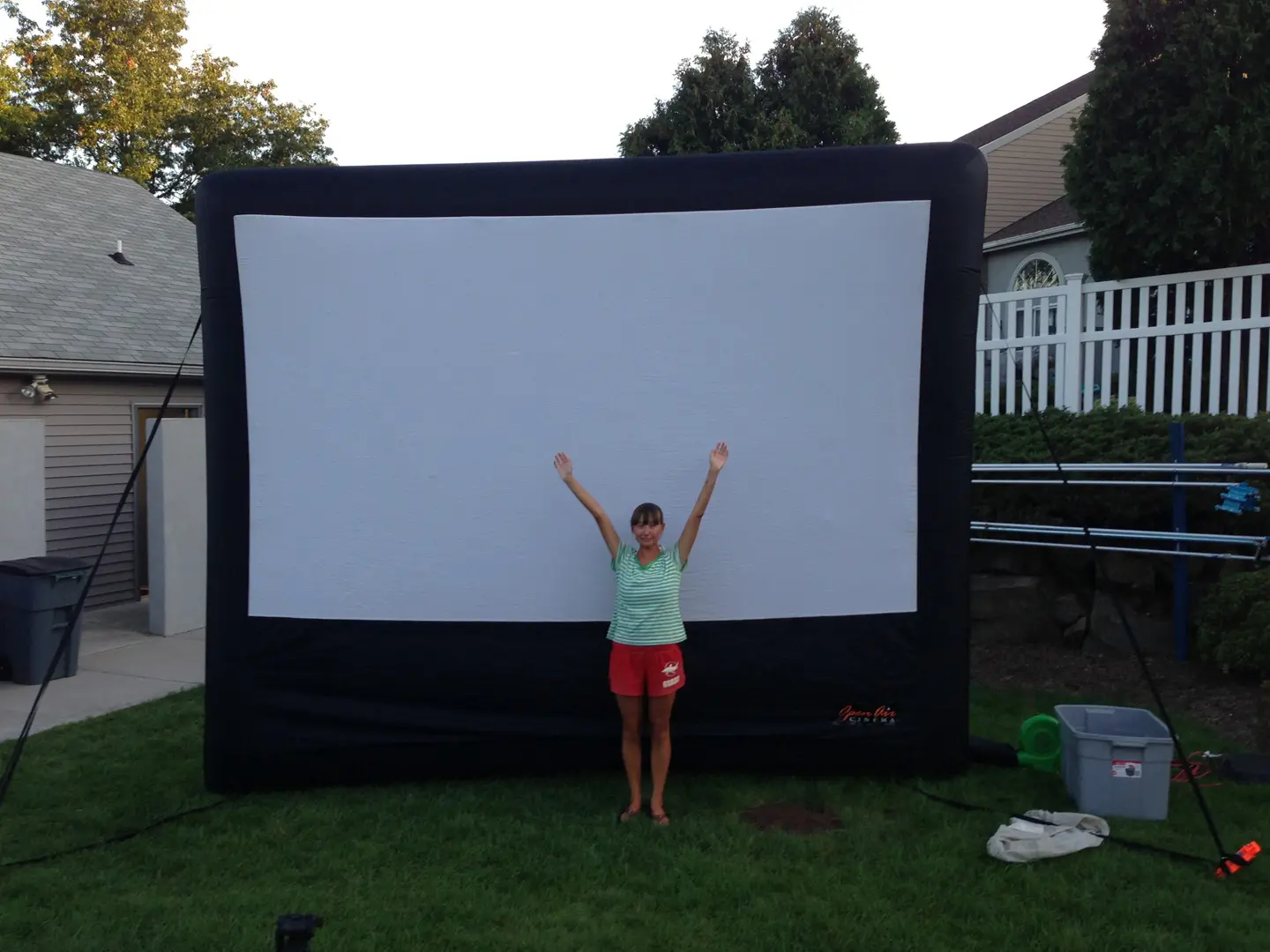 A person stands with arms raised in front of a large inflatable movie screen in a backyard, surrounded by houses and trees.