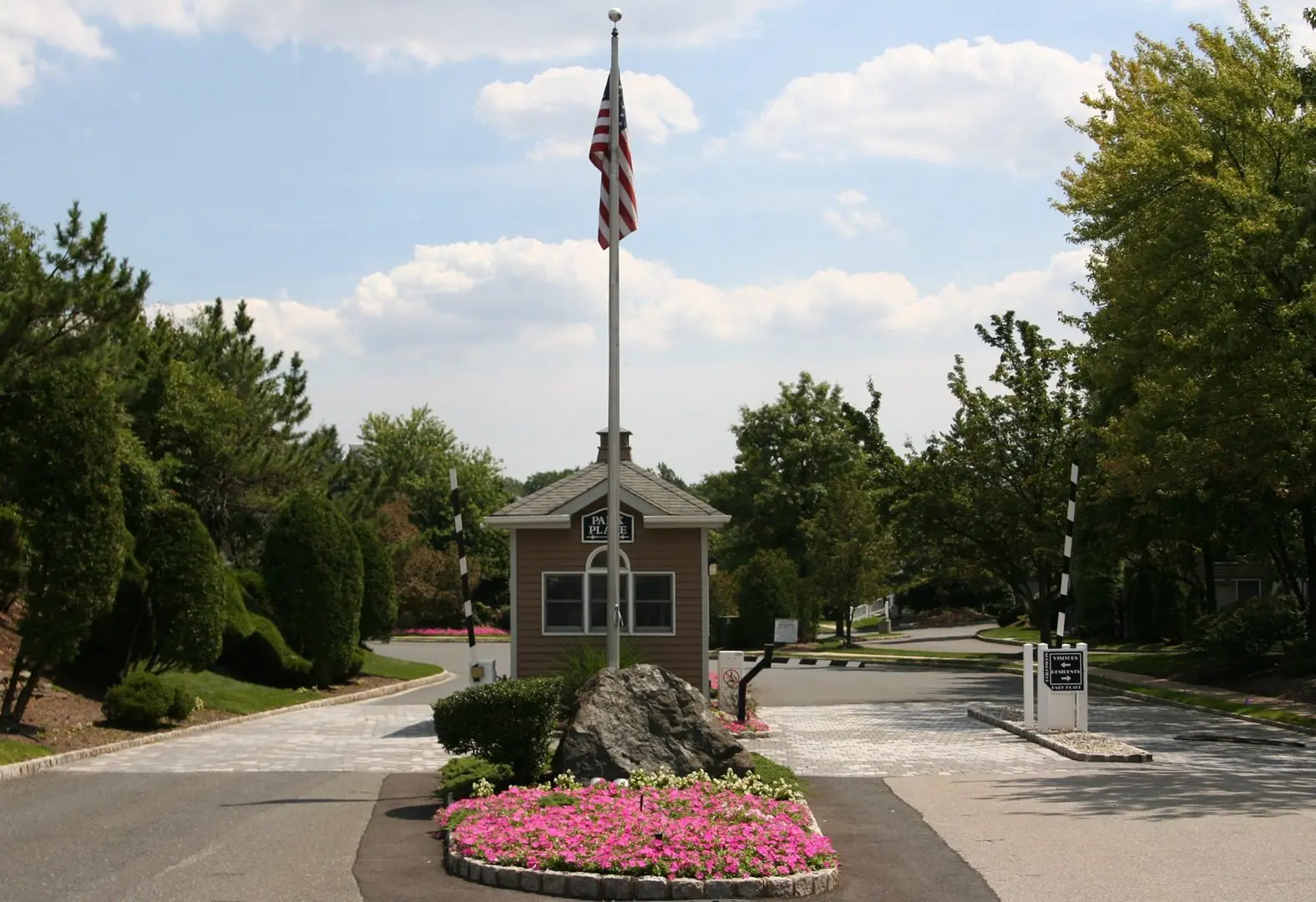 A small guardhouse with an American flag stands in the center of a driveway. Flowers are planted in front, and trees line the road.