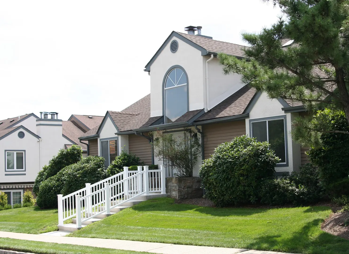 A suburban house with a front porch, arched window, and sloped roof, surrounded by green shrubs and a lawn, features a white ramp leading to the entrance.