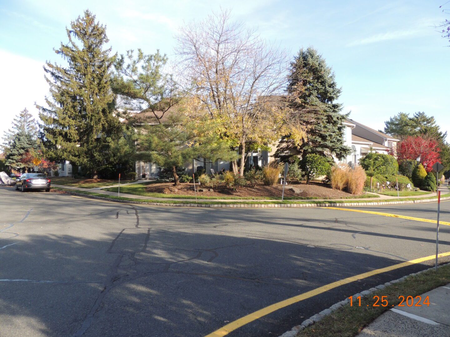 Residential street corner with houses, trees, and clear skies. Cars parked along the roadside, date stamp on bottom right reading 11-25-2024.