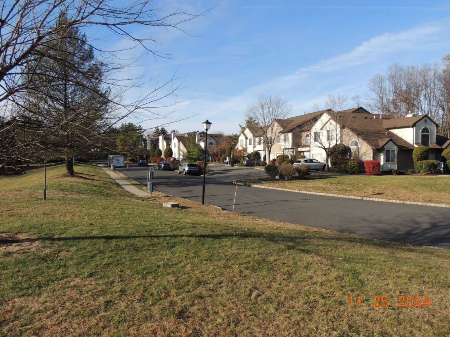Suburban neighborhood street on a clear day with houses and trees lining the road. Grass and sidewalks are visible.