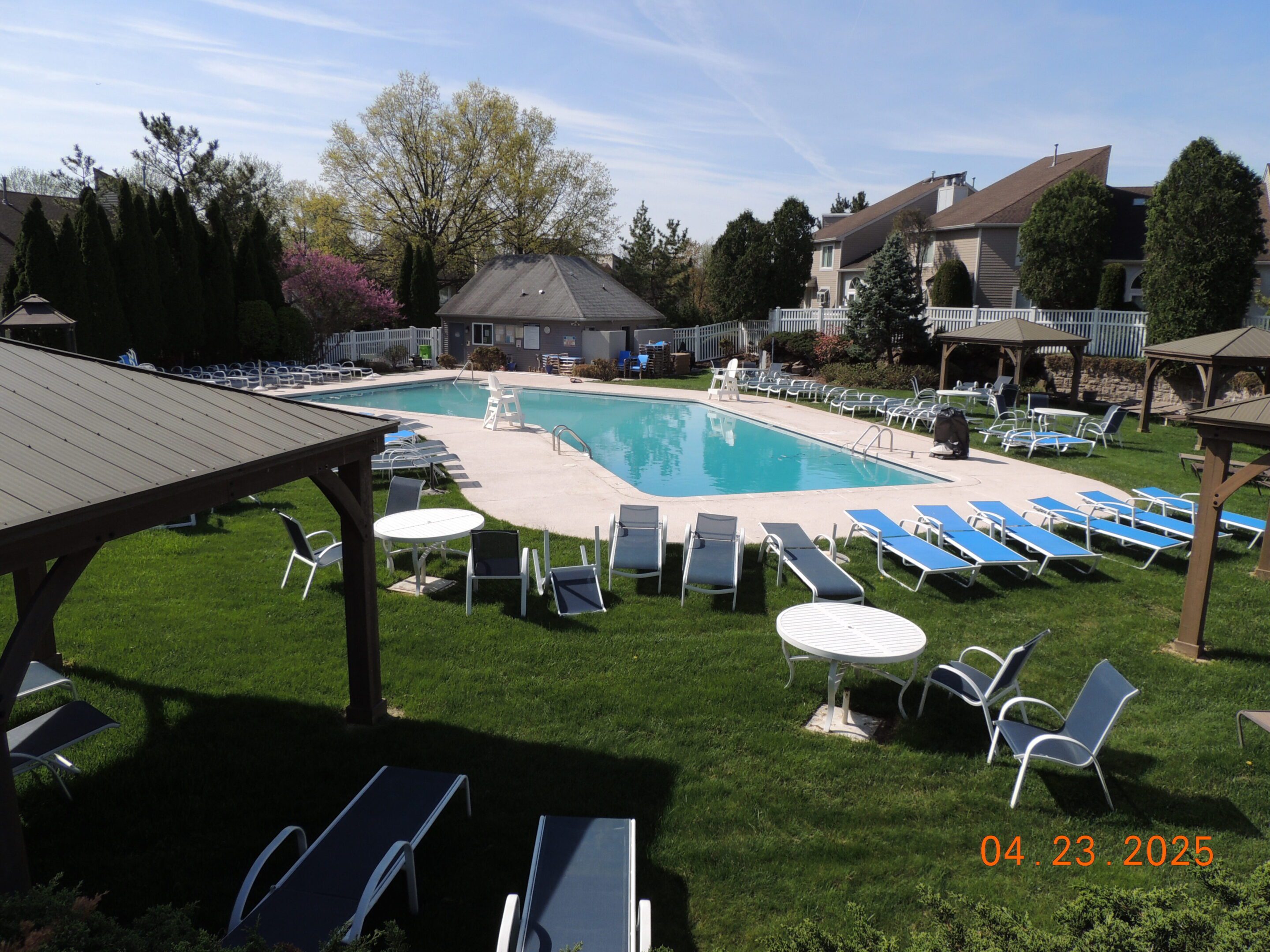 Outdoor swimming pool surrounded by empty lounge chairs and tables on a sunny day with residential buildings in the background.
