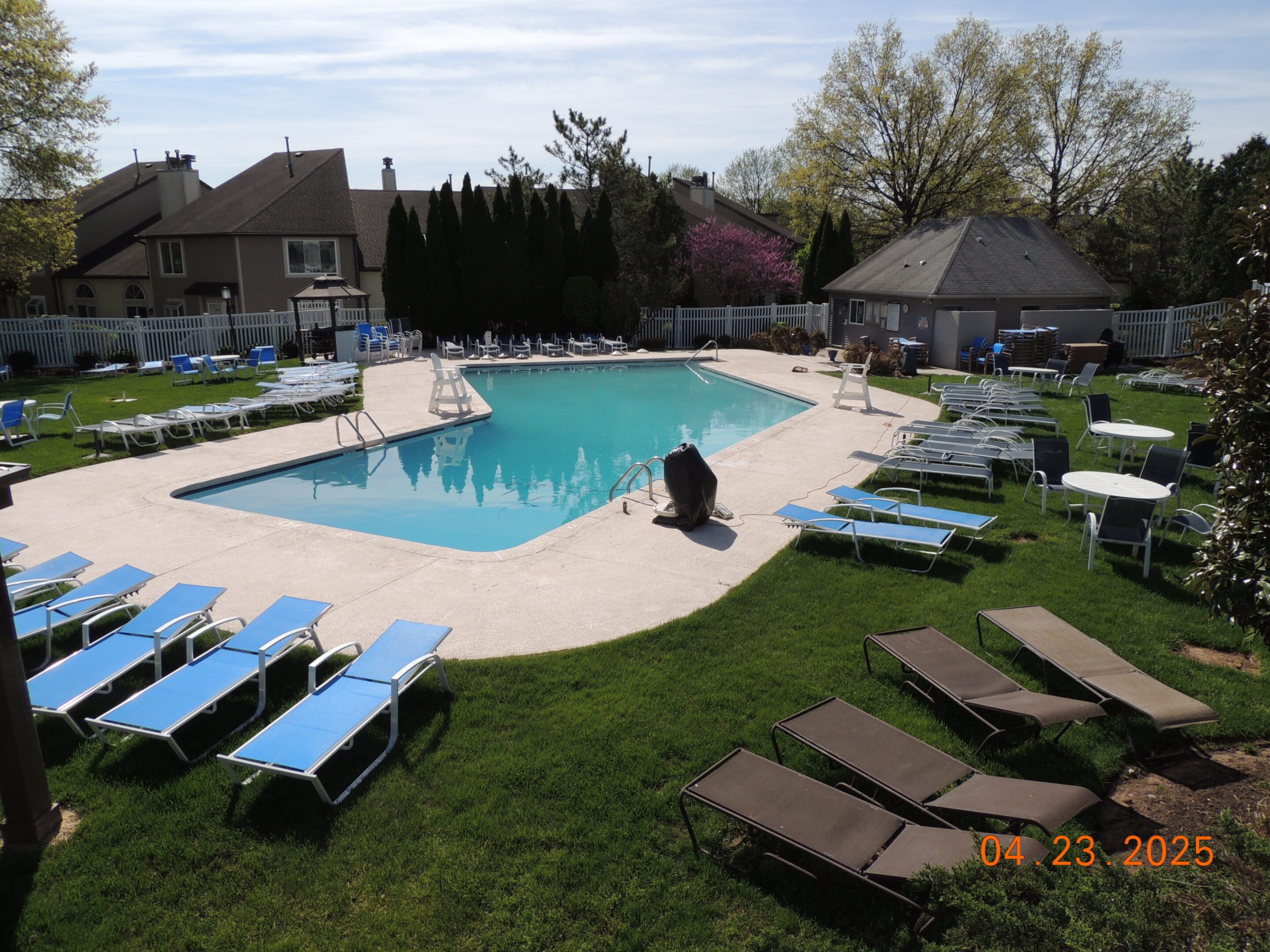 Outdoor swimming pool surrounded by lounge chairs and tables on a grassy area, with residential buildings and trees in the background. Photo taken on April 23, 2025.