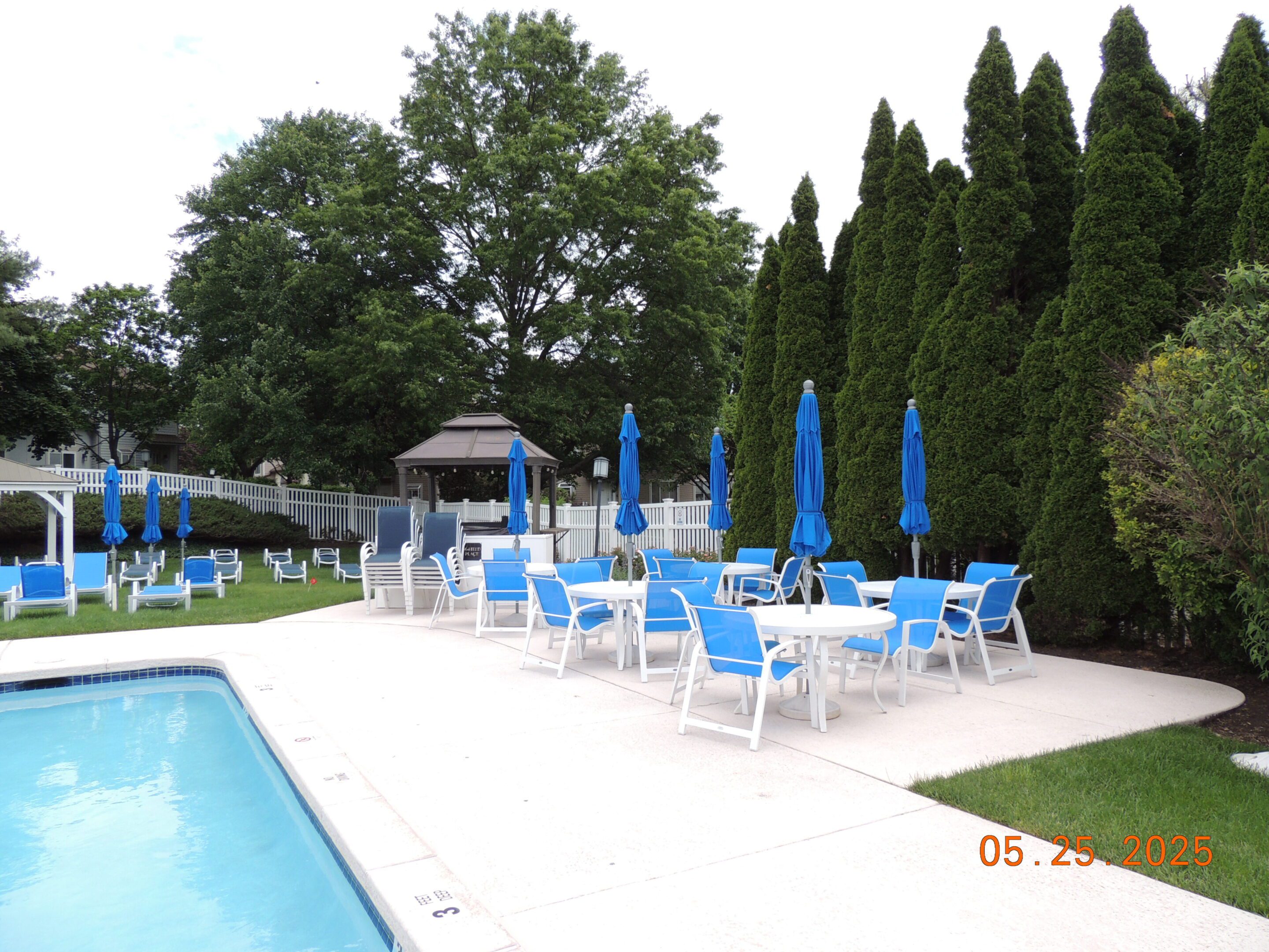 Outdoor poolside area with blue and white lounge chairs, tables, and closed umbrellas, surrounded by trees and greenery on a cloudy day.