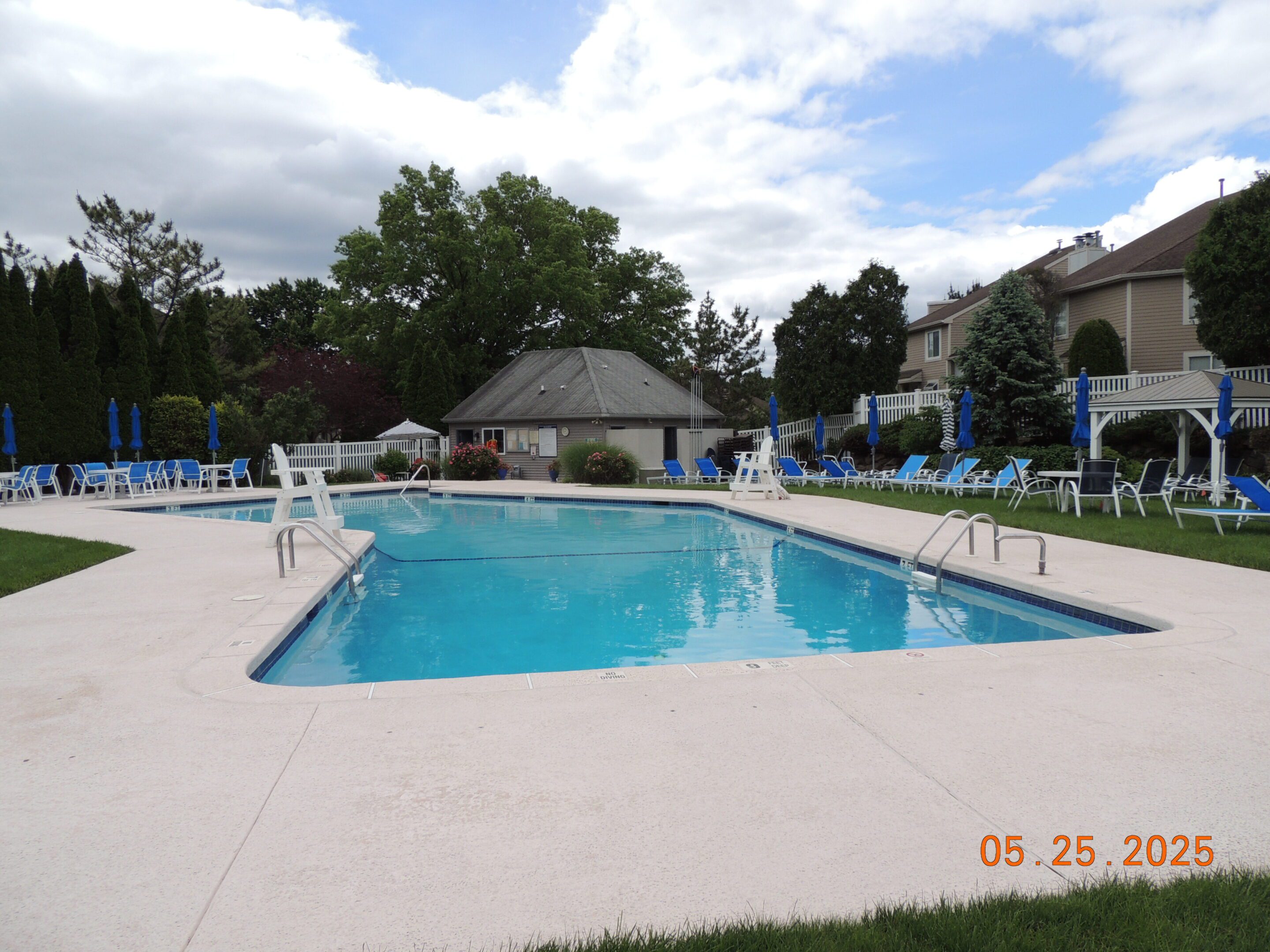 Rectangular outdoor swimming pool with blue umbrellas and lounge chairs around it, trees and houses in the background, dated May 25, 2025.