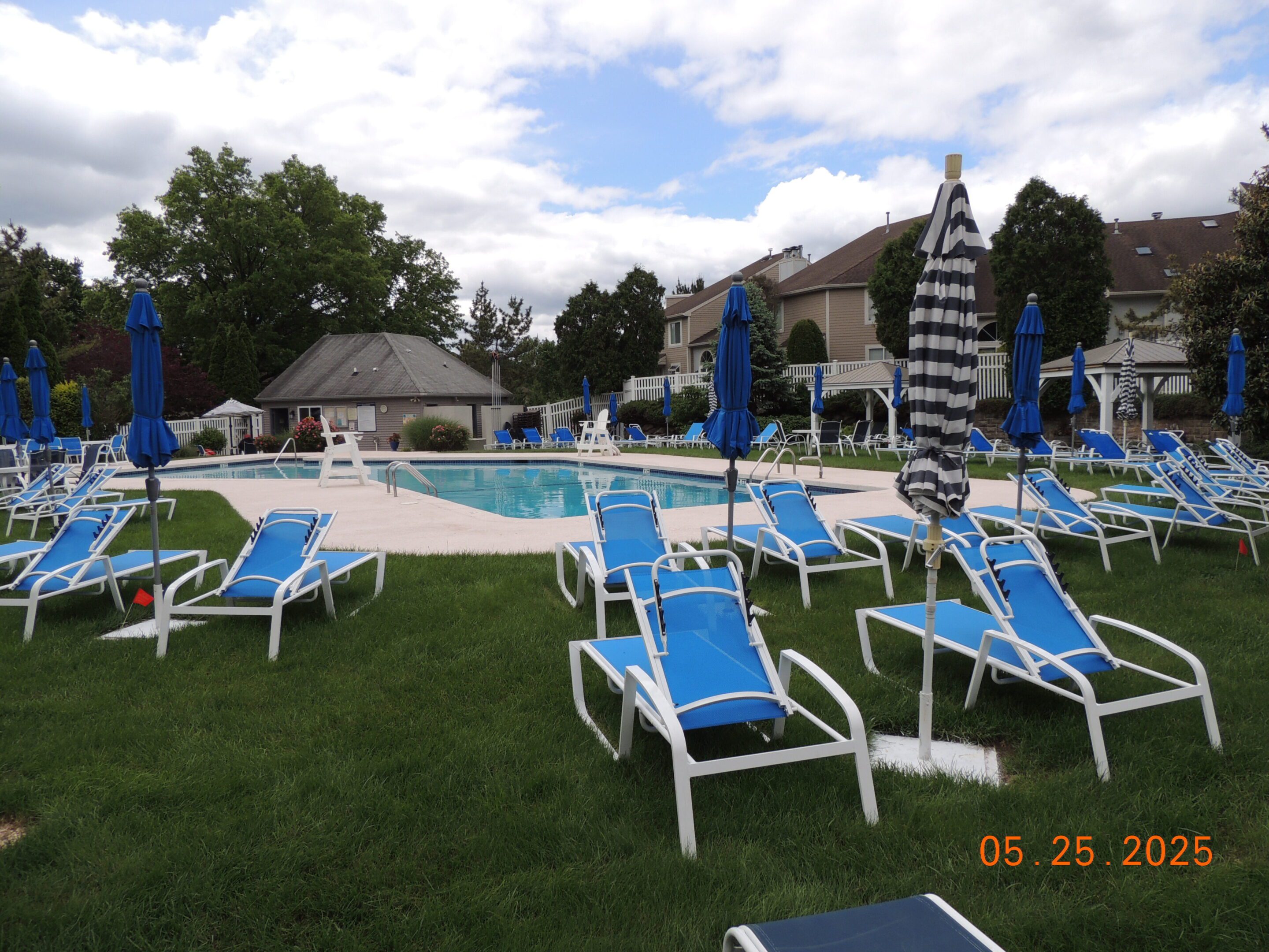 Outdoor swimming pool surrounded by blue lounge chairs and closed umbrellas on a cloudy day, with houses and trees in the background.