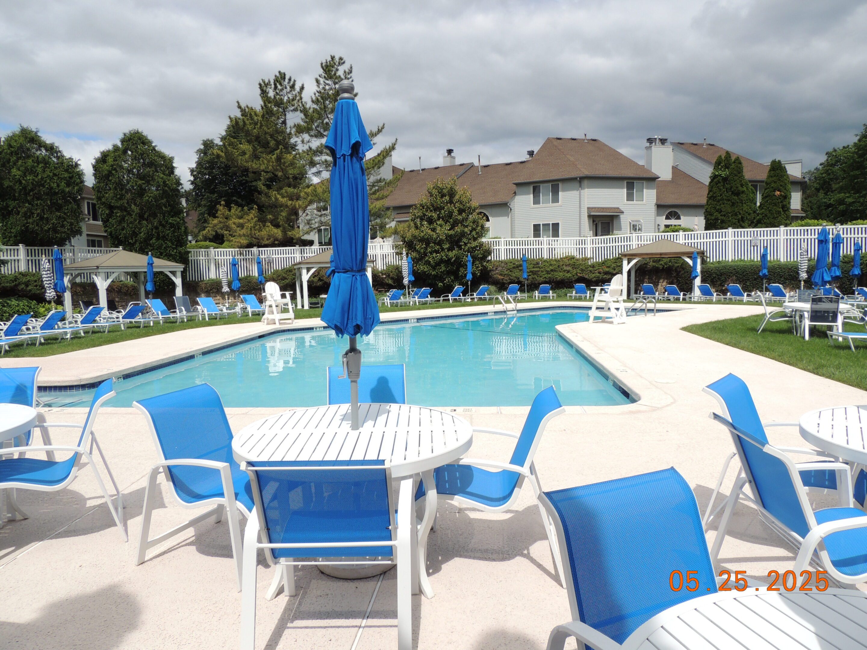 Outdoor swimming pool surrounded by blue and white lounge chairs and tables, with patio umbrellas and residential buildings in the background.