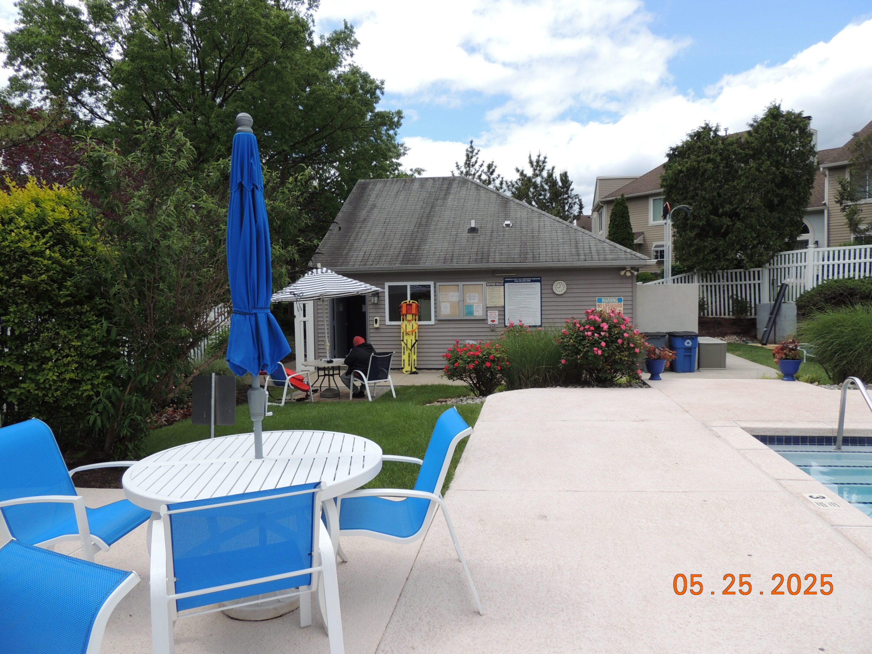 Outdoor poolside area with blue and white chairs and tables, a closed umbrella, pool edge, and a small building in the background. Date stamp reads "05. 25. 2025.
