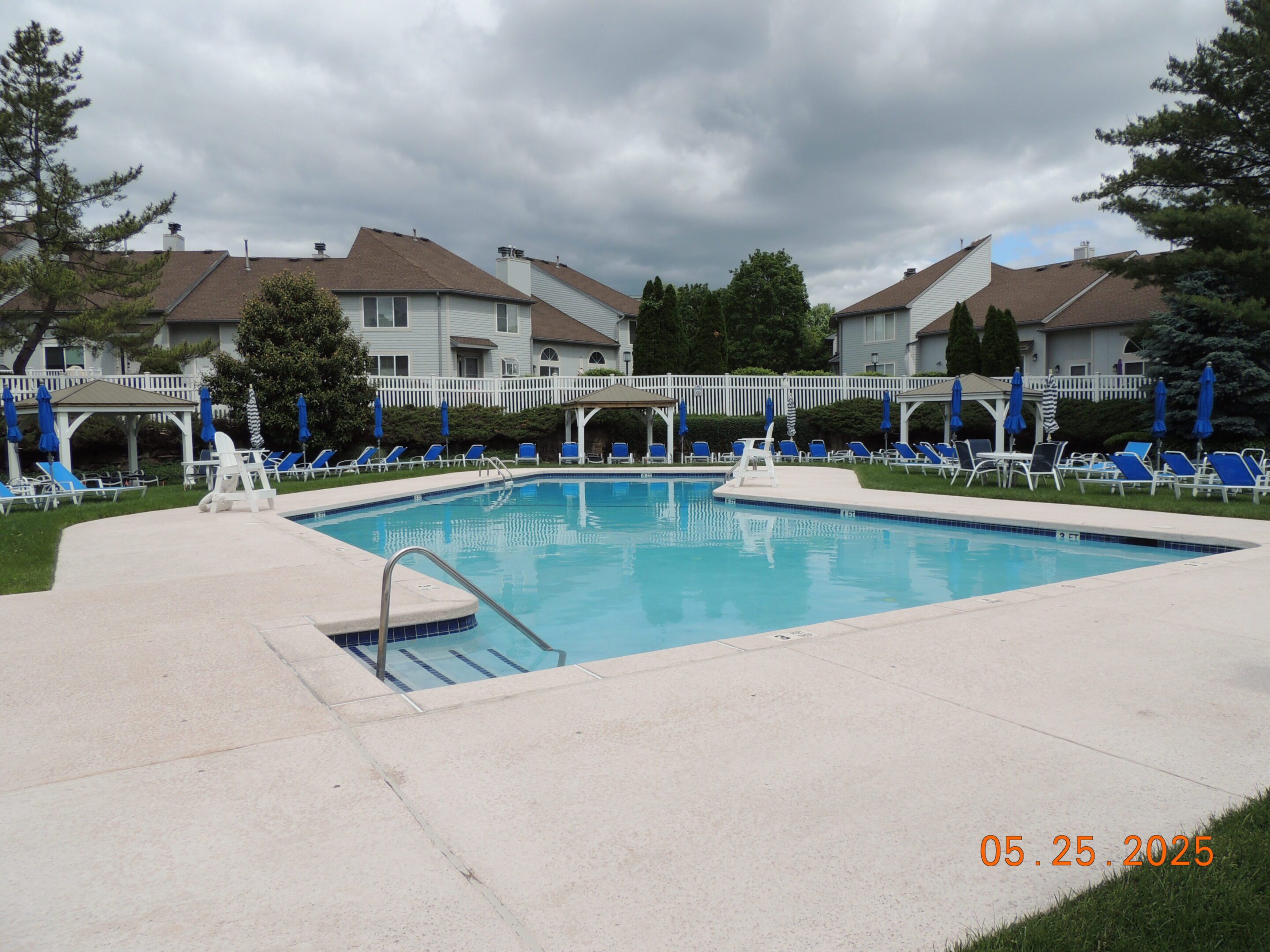 Outdoor swimming pool surrounded by blue lounge chairs and umbrellas in a residential townhouse complex, under a cloudy sky. Dated May 25, 2025.