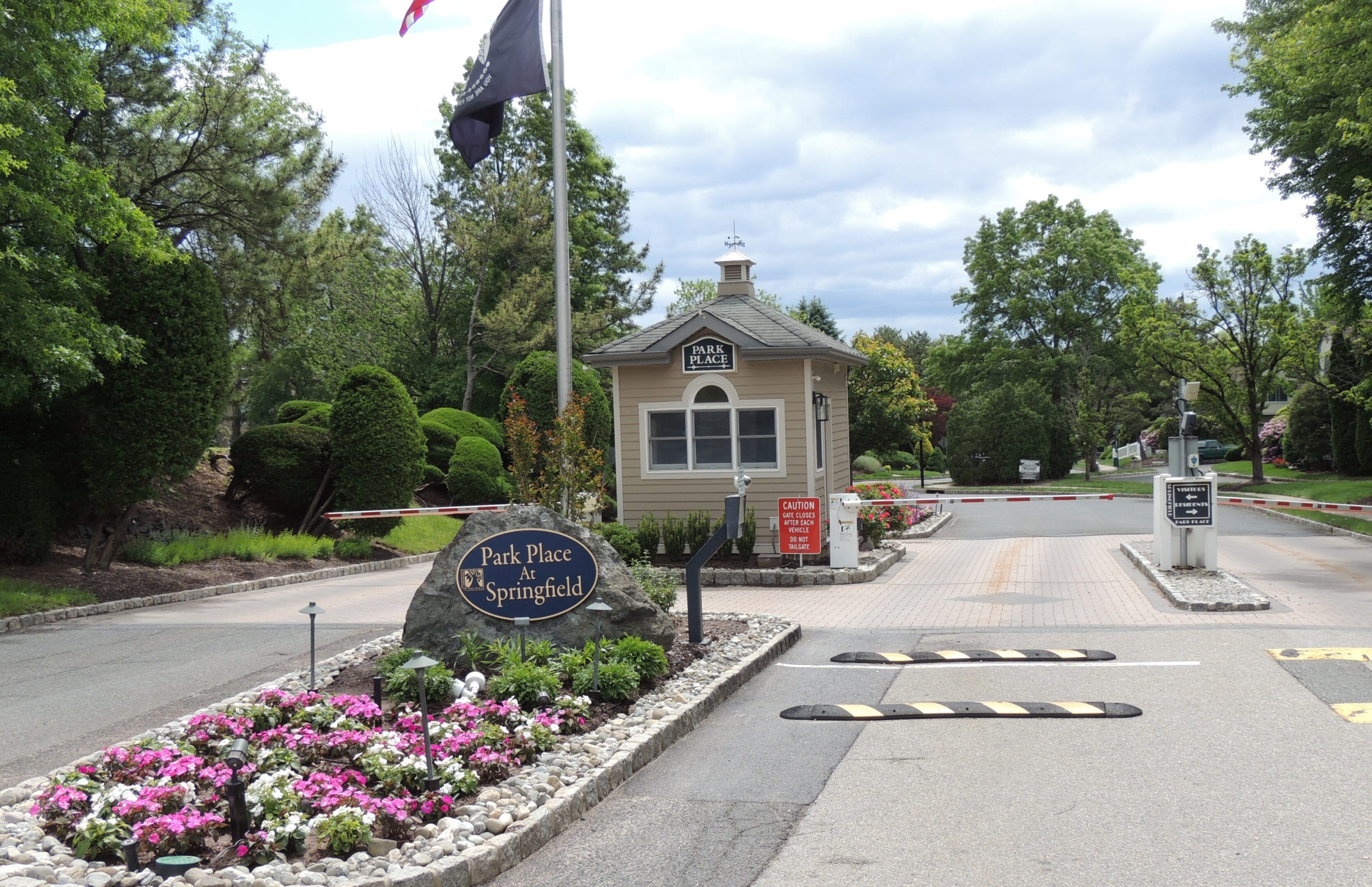 Entrance gate to Park Place at Springfield, featuring a guard booth, landscaped flower bed, and security barriers on a driveway under a cloudy sky.