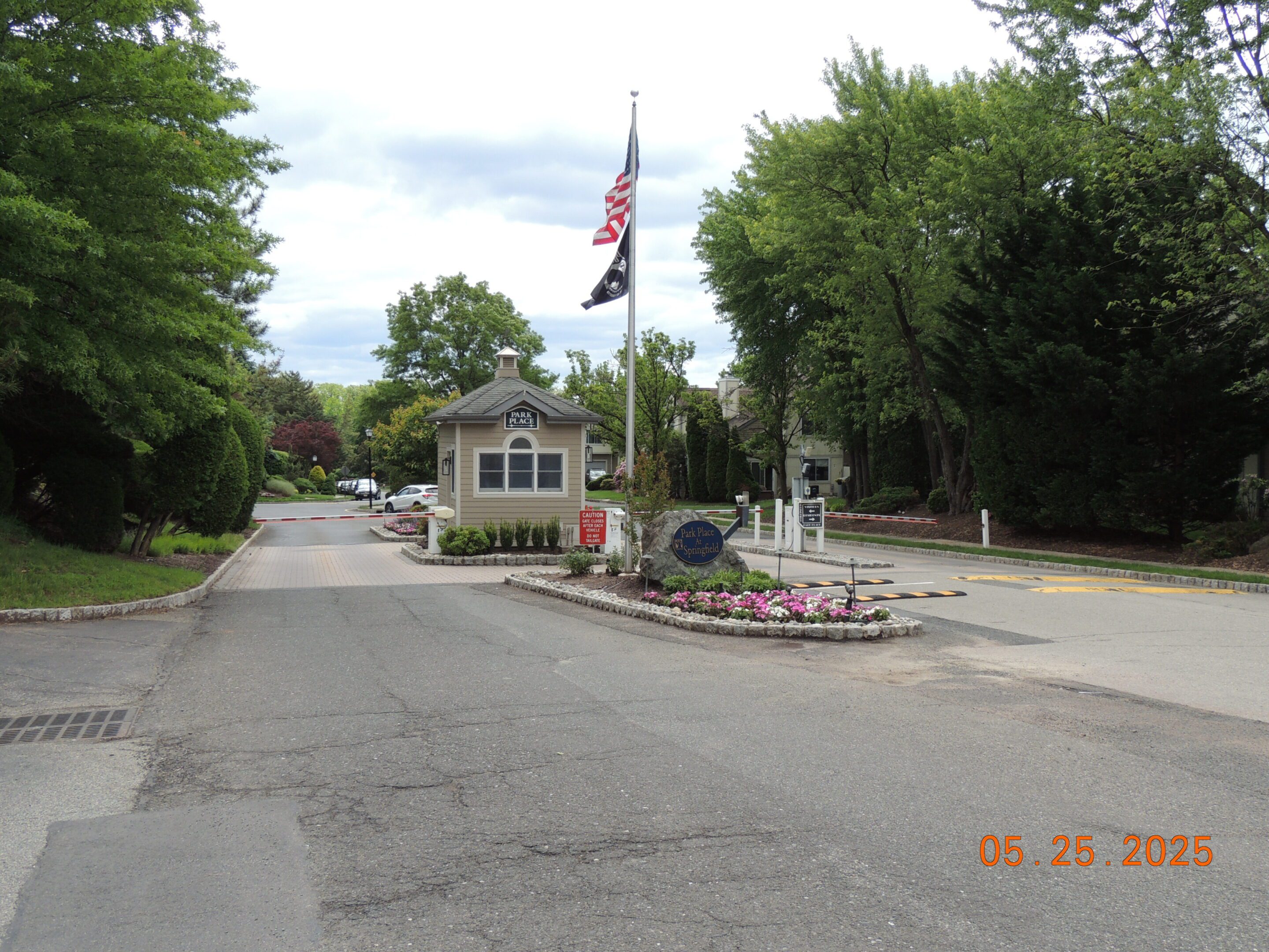 A gated community entrance features a small guardhouse, flagpole with US and POW/MIA flags, flower bed, and security barriers. Photo is dated May 25, 2025.