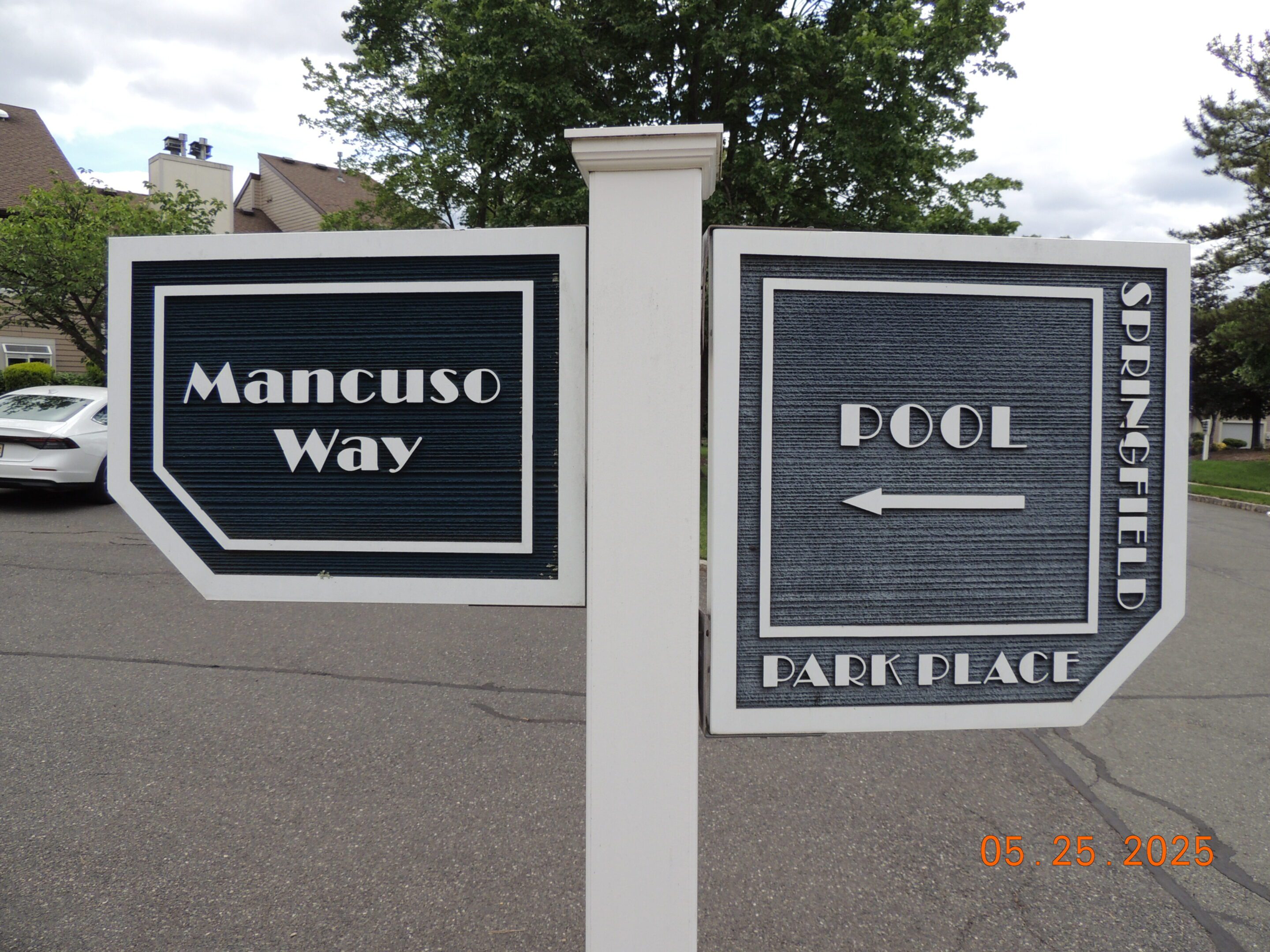 Two directional signs: one points left labeled "Mancuso Way," the other points right labeled "Pool" with an arrow, "Springfield," and "Park Place." Trees and a parked car in background.