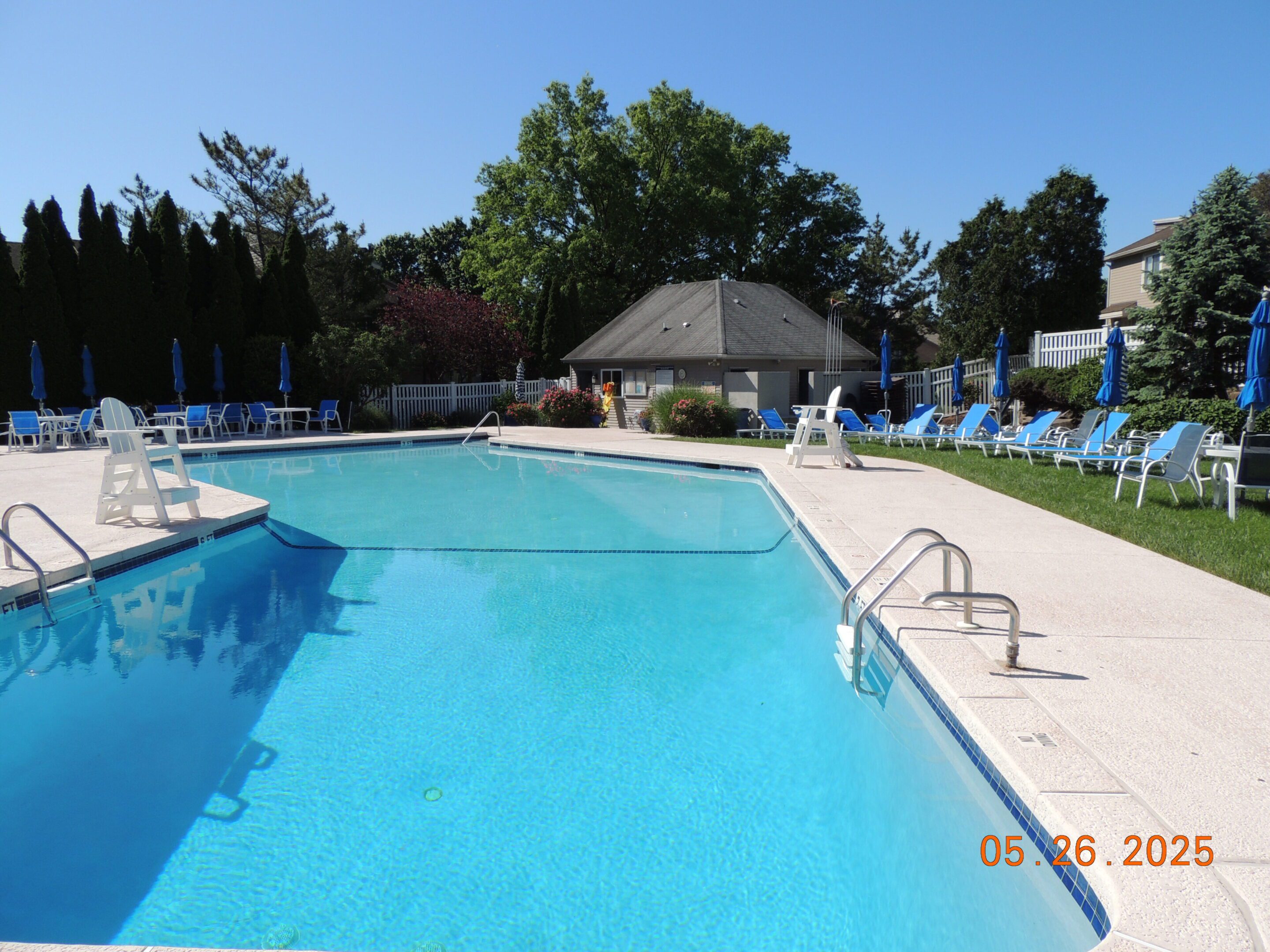 Outdoor swimming pool with clear blue water, surrounded by lounge chairs and umbrellas, with a small building in the background; date stamp reads 05.26.2025.
