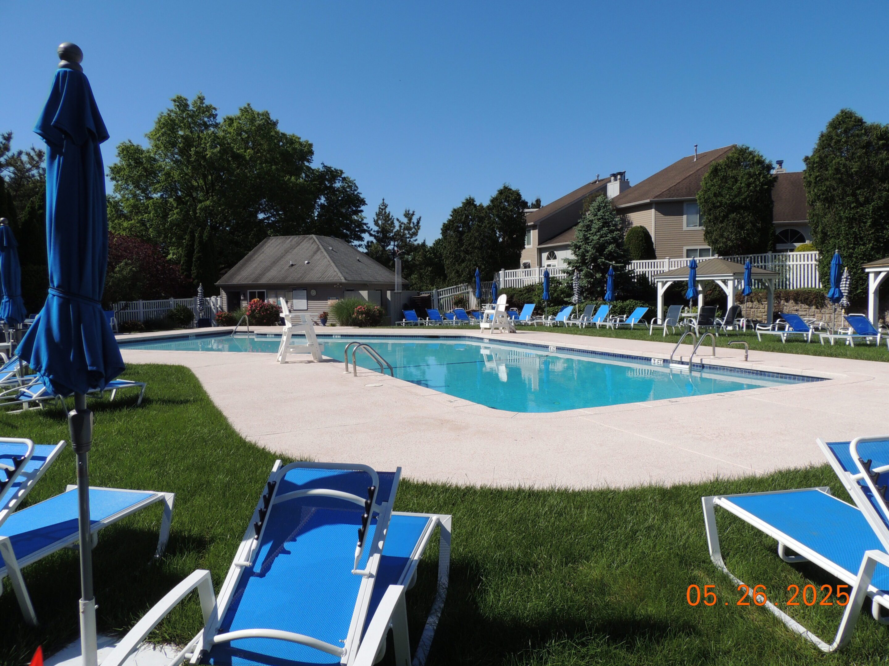 Outdoor swimming pool surrounded by blue lounge chairs and umbrellas on a sunny day, with trees and houses in the background.
