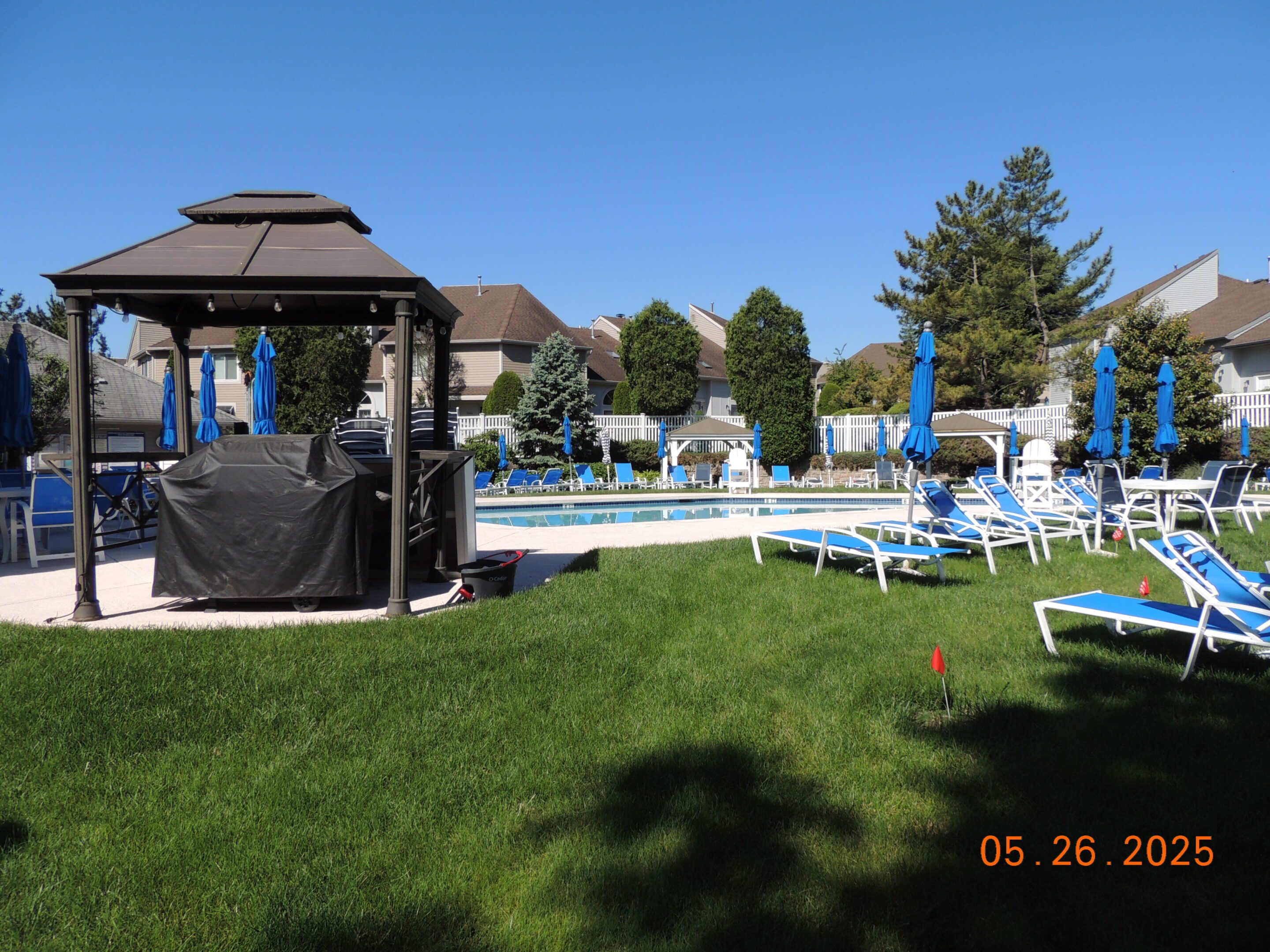 Outdoor pool area with blue lounge chairs, umbrellas, a gazebo, and surrounding buildings on a sunny day. Date on photo: May 26, 2025.
