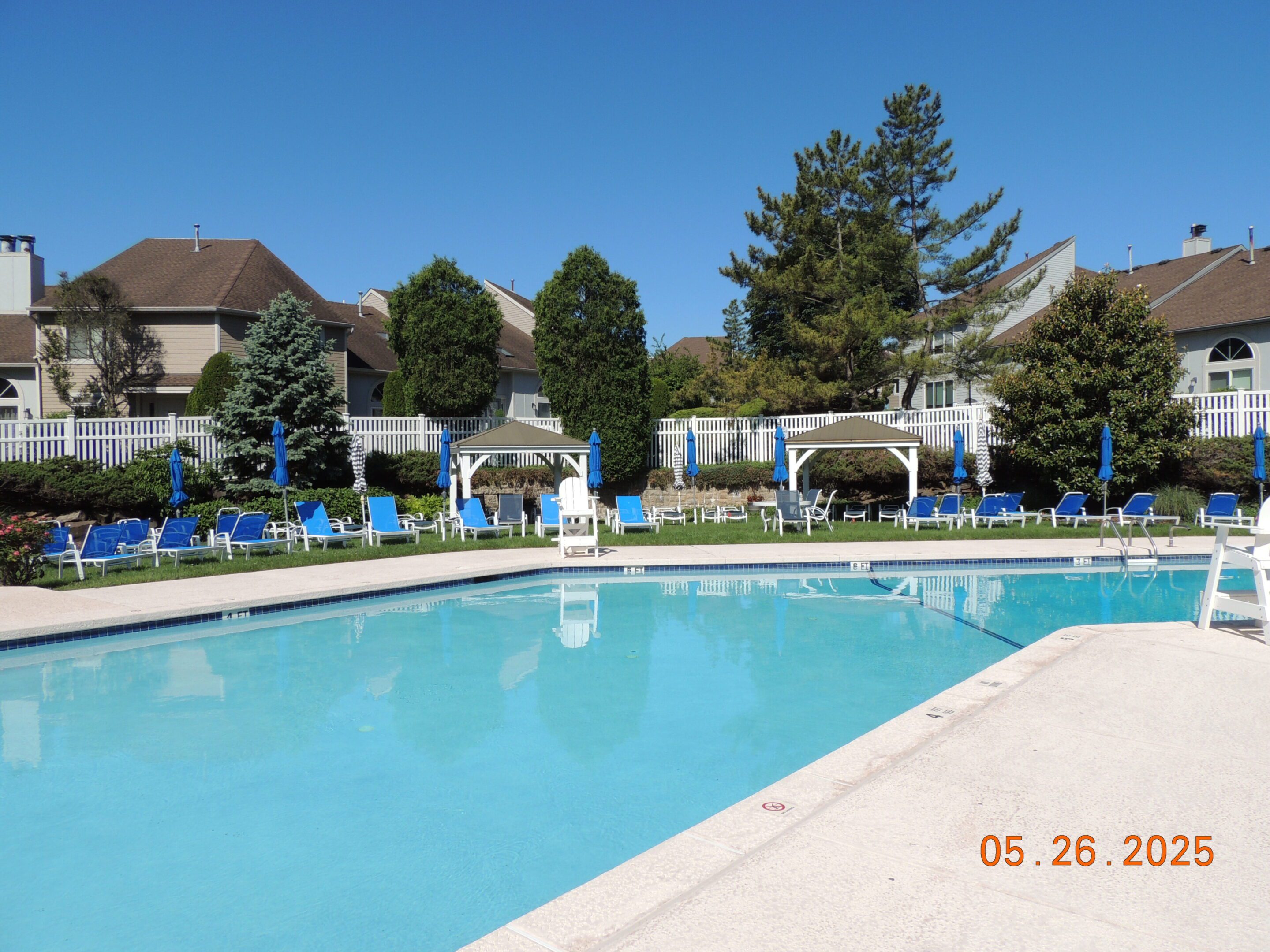 Outdoor swimming pool surrounded by blue lounge chairs and umbrellas, with trees and residential buildings in the background under a clear blue sky. Date on image: May 26, 2025.
