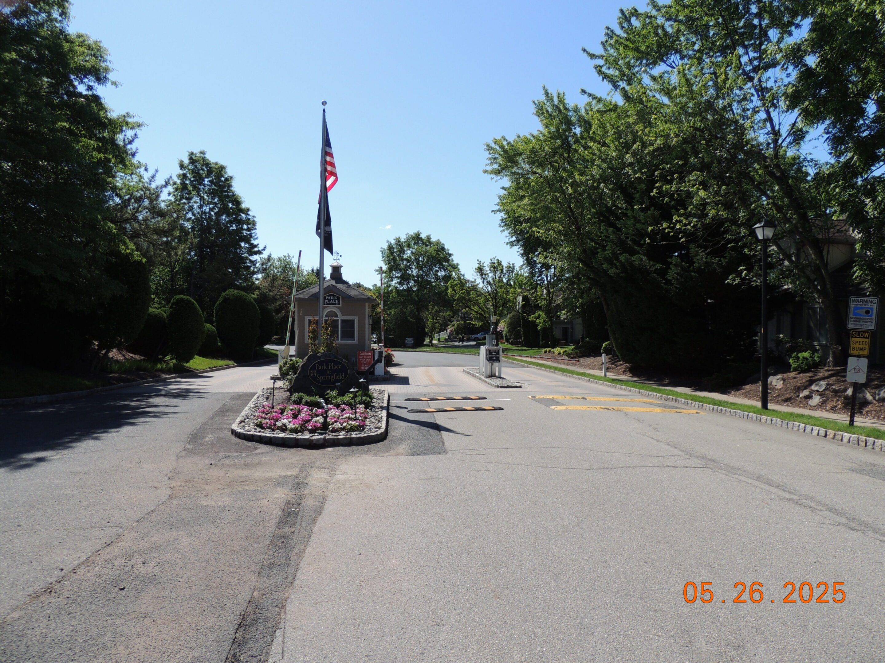 A gated entrance with an American flag, a small guardhouse, flower bed, and speed bumps under clear skies. Date stamp: May 26, 2025.